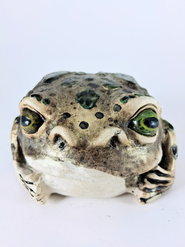 A close-up photograph of the front of a frog sculpture made of raku clay. The sculpture is very round and brown with dark green and black speckles. It has green eyes.